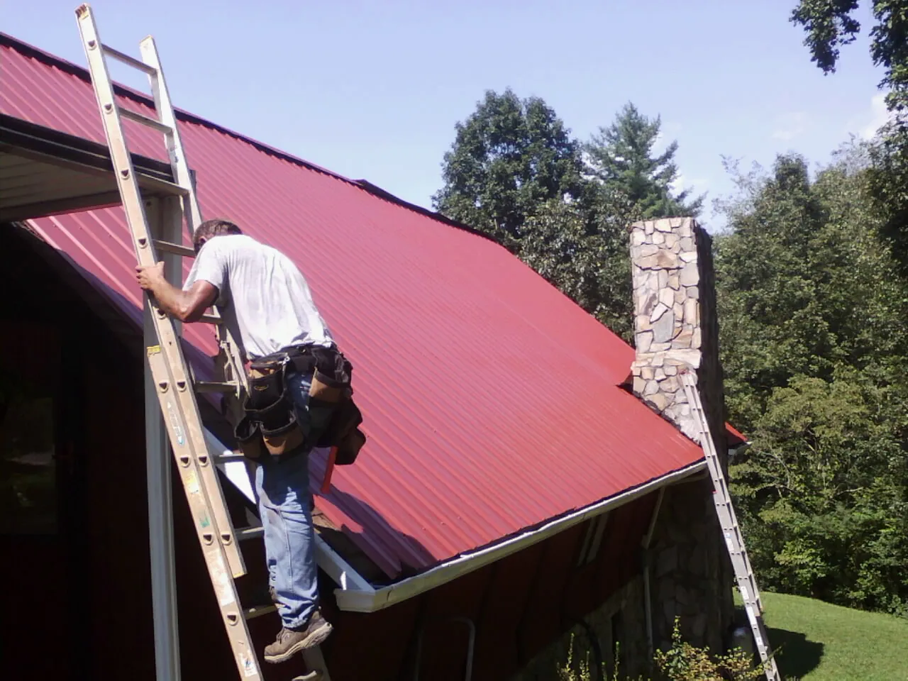 Roofer on a ladder beside a red metal roof and stone chimney — Soffit & Fascia Repair by Roofing Brothers in Mays Landing